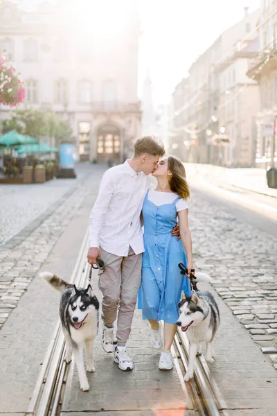 Couple in love with husky dogs, kissing and walking on city street. Beautiful view of old European city, summer sunny day and ancient buildings on the background — Stock Photo, Image