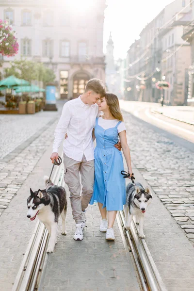 Casal romântico na cidade com seus dois cães husky, andando na trilha do bonde na estrada do pavimento, abraçando e olhando um ao outro. Jovem senhora em vestido azul e homem atraente com cães na cidade — Fotografia de Stock