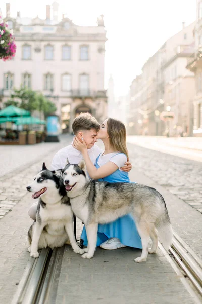 Happy cheerful young couple with two cute husky dogs walking in the city in summer sunny day. Charming young girl in blue dress kissing forehead of her handsome boyfriend — Stock Photo, Image