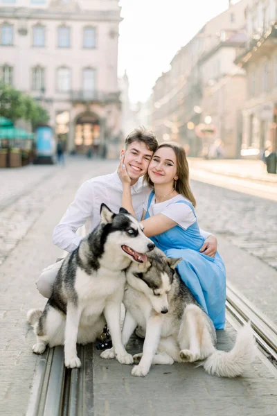 Happy romantic young couple posing to camera with smile, while sitting on the street in old European city outdoors, walking with their adorable gray husky dogs — Stock Photo, Image