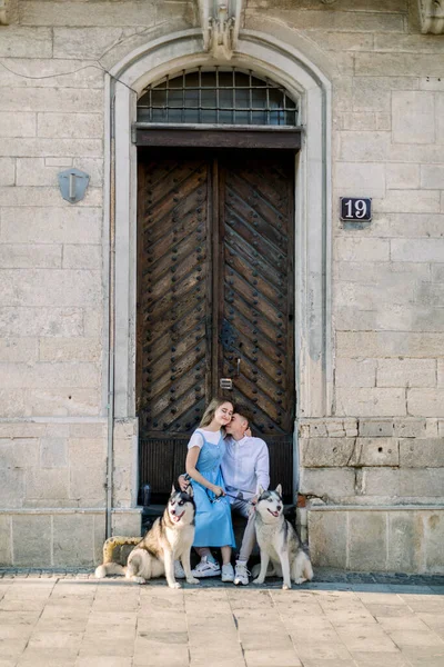 Gelukkig stel met twee husky honden die plezier hebben buiten in de stad, zitten in de buurt van oude houten deur in de voorkant van het oude stenen gebouw. Volledige foto — Stockfoto