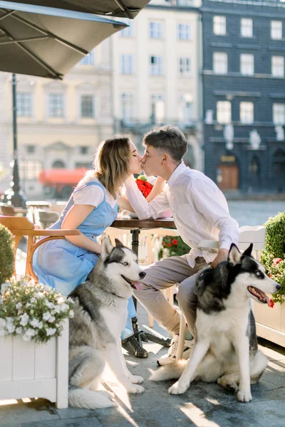 Mooi jong paar in liefde genieten van zomerwandeling in de stad straat met hun husky honden, zitten aan de tafel in cafe buiten en zoenen — Stockfoto