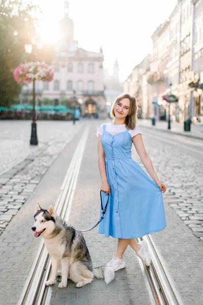 Pieno ritratto estivo all'aperto di giovane ragazza attraente in abito blu, in posa con il suo cane husky, in piedi sulla pista del tram sullo sfondo di vecchi edifici nella città europea — Foto Stock