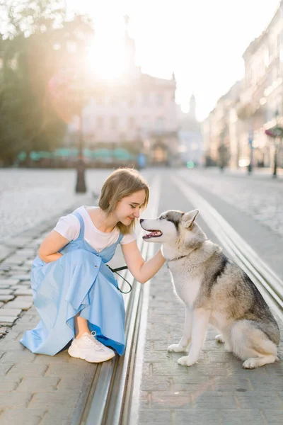 Молода і вродлива жінка у блакитній сукні бавиться зі своїм гарненьким собакою у сонячний ранок у старому європейському місті. — стокове фото