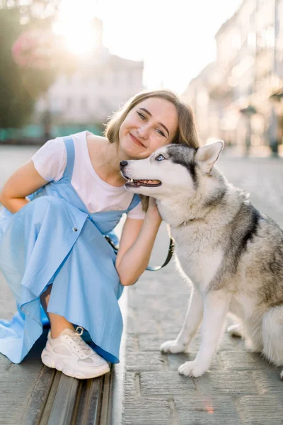 Cerca de retrato al aire libre de la hermosa joven, con vestido azul casual y zapatillas de deporte, sentado en el camino del pavimento y abrazando a su lindo perro husky en la ciudad, durante el día de verano soleado —  Fotos de Stock