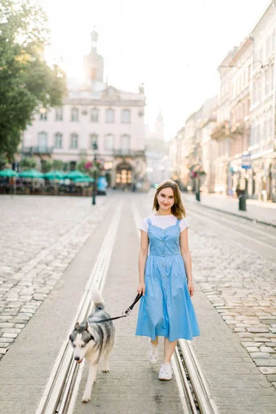 Pieno ritratto estivo all'aperto di giovane ragazza attraente in abito blu, in posa con il suo cane husky, in piedi sulla pista del tram sullo sfondo di vecchi edifici nella città europea — Foto Stock