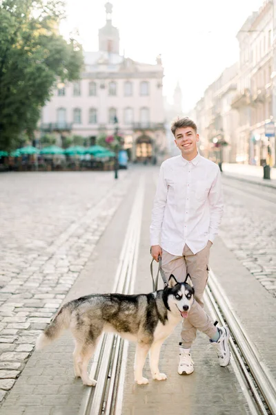 Morning walk with husky dog in the city. Young handsome hipster man walking with his husky dog on the old European city street at golden sunrise — Stock Photo, Image