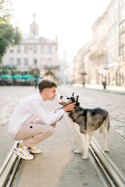 Міська прогулянка з собакою. Молодий красивий хлопчик у повсякденному одязі, гуляючи зі своїм гамірним собакою по дорозі на тротуарі і трамвайною колією на старій європейській міській вулиці на світанку. — стокове фото