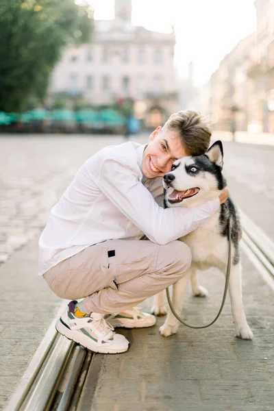 Концепція домашніх тварин, собак і людей. Щасливий усміхнений молодий чоловік обіймає милого хаскі собаку, гуляючи по міській вулиці в сонячний літній день — стокове фото
