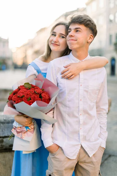Happy young couple in love, enjoying their walk in beautiful ancient city, posing to camera near old stone fountain. Pretty girl holds red flowers and hugs her boyfriend from behind — Stock Photo, Image