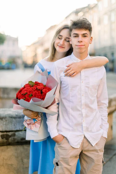 Happy young couple in love, enjoying their walk in beautiful ancient city, posing to camera near old stone fountain. Pretty girl holds red flowers and hugs her boyfriend from behind — Stock Photo, Image