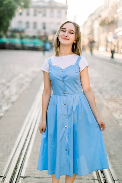 Tiro de menina bonita jovem em vestido azul elegante, posando na bela rua, de pé na pista de bonde e estrada de pavimento velho, na cidade antiga ao pôr do sol. — Fotografia de Stock