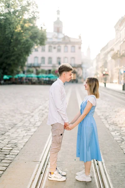 Happy to be together. Portrait of beautiful young couple holding hands and looking at each other with smile while standing on the tram track on pavement road in old European city — Stock Photo, Image