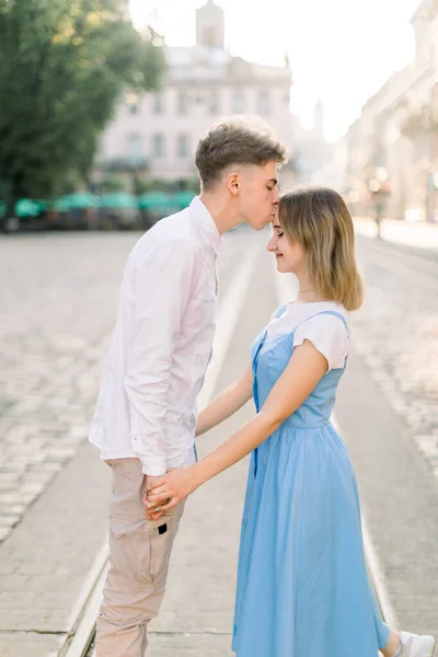 Handsome young man kissing his charming pretty girlfriend on forehead in the street in the sunny morning. Romantic couple in the street in ancient European city — Stock Photo, Image