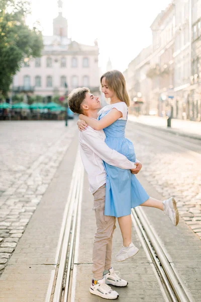 Couple adorable amoureux, profitant de leur promenade par une journée ensoleillée dans la ville, debout sur la piste de tram. beau gars tient son attrayant jeune fille en robe bleue sur les mains — Photo