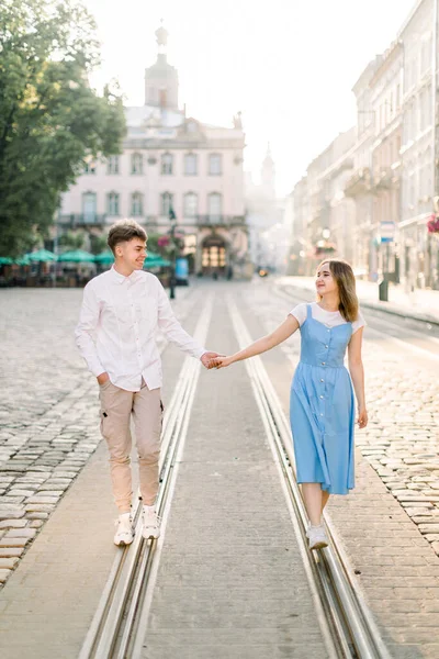 Feliz joven pareja con estilo, chica en vestido azul y hombre guapo disfrutando de su paseo, divirtiéndose en la pista de tranvía en el camino viejo pavimento en la calle de la ciudad con el edificio antiguo en el fondo. —  Fotos de Stock