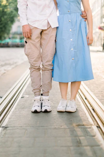 Image recadrée des jambes d'un beau jeune couple élégant amoureux, femme en robe bleue et homme en pantalon beige, debout sur la chaussée et la piste de tramway, s'amusant en plein air dans la ville — Photo