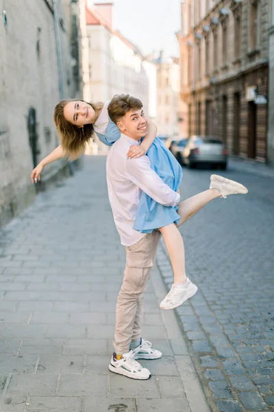 Happy couple having fun outdoors in the city. Young handsome man holding his lovely girlfriend in blue dress in his arms outdoors, walking in the street — Stock Photo, Image