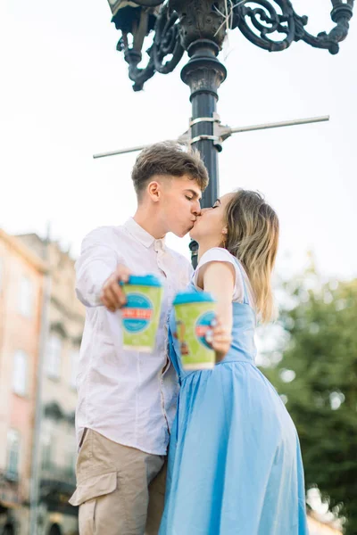 Close up retrato de romântico beijando jovem casal hipster no amor, mostrando copos de tomar café para câmera, de pé sob a luz da rua na cidade antiga ao ar livre — Fotografia de Stock