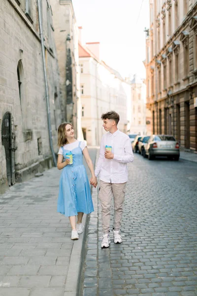 Viajes pareja de turistas caminando por la calle. Retrato de la hermosa joven en vestido azul y su novio guapo en ropa elegante turismo atracciones de la ciudad, con tazas de café para llevar —  Fotos de Stock