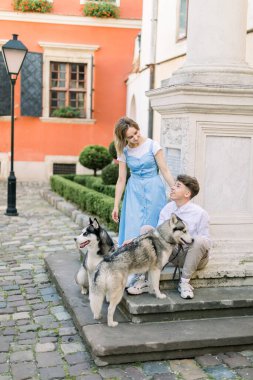 İki köpekli mutlu çift şehirde dışarıda eğleniyor. Bahçedeki anıtın yanındaki merdivenlerde oturuyorlar. Arka planda turuncu bina var.