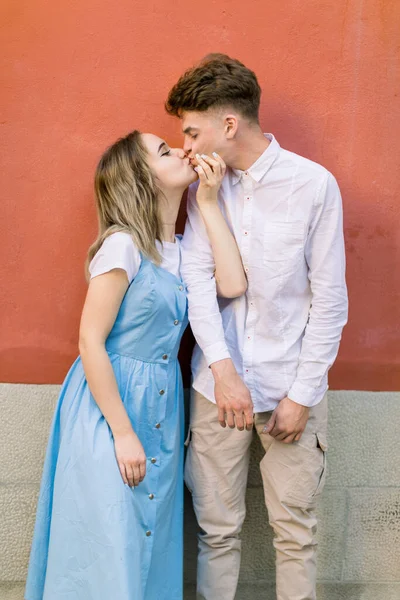 Casal, relações, retrato ao ar livre. Elegante hipster casal feliz, cara na camisa branca e menina em vestido azul, se divertindo ao ar livre na cidade, de pé na frente da parede laranja e beijando — Fotografia de Stock