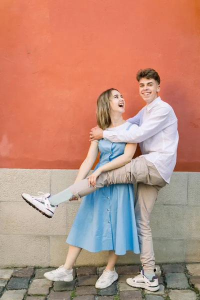 Happy crazy funny couple in summer casual clothes, having fun outdoors in the city, posing to camera, laughing and raising their legs in sneakers up