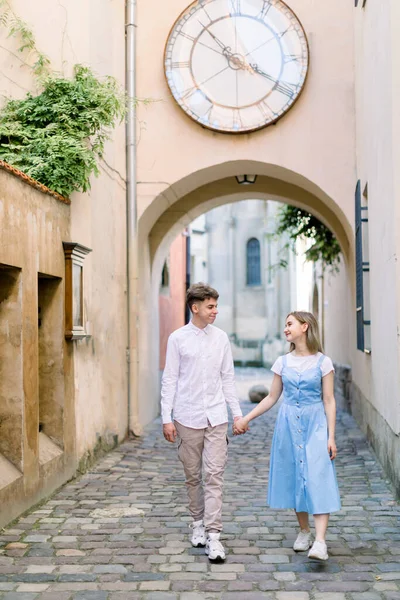 Retrato al aire libre de la ciudad de feliz pareja enamorada, novio guapo y chica joven atractiva en vestido azul, caminando juntos en la hermosa ciudad europea, tomados de la mano y sonriendo —  Fotos de Stock