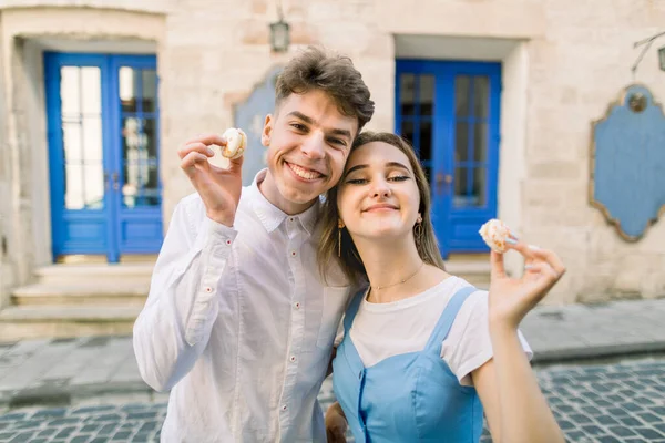 Horizontal shot of funny lovely young couple, walking outdoors in the city, posing to camera with tasty macarons on the background of vintage building with blue door — Stock Photo, Image