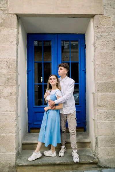 Preciosa cita en la calle. Feliz pareja joven hipster, hombre guapo con su atractiva novia en vestido azul, abrazándose al aire libre en la ciudad, posando cerca de la hermosa puerta azul vintage —  Fotos de Stock