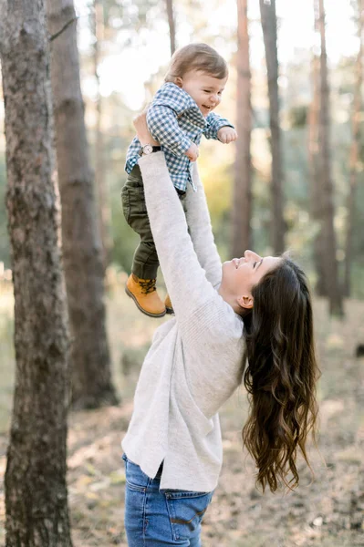 Portrait of happy Caucasian mother lifting and playing with her cute little son, enjoying their joint walk in pine forest together. Happy family concept, mom and son in forest — Stock Photo, Image