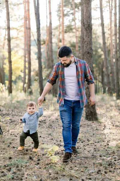 Retrato ao ar livre de comprimento total de belo pai jovem barbudo, andando na floresta de outono de pinho, juntamente com seu pequeno filho adorável — Fotografia de Stock