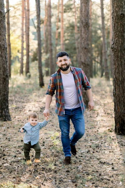Retrato ao ar livre de comprimento total de belo pai jovem barbudo, andando na floresta de outono de pinho, juntamente com seu pequeno filho adorável — Fotografia de Stock