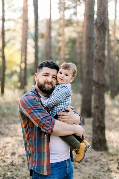 Feliz jovem pai barbudo segurando seu filho, garoto engraçado, olhando para a câmera enquanto caminham juntos em um parque ou floresta no dia ensolarado de outono. Família feliz, alegre, pai e filho — Fotografia de Stock