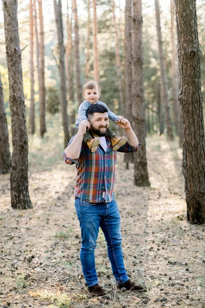 Jovem bonito pai e filho, vestindo roupas casuais elegantes, se divertindo no parque de outono. Explorando a natureza com o pai no outono da floresta, o pai segura seu filho nos ombros — Fotografia de Stock