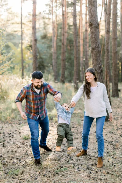 Mutlu mutlu aile, yakışıklı sakallı baba, güzel esmer anne ve sevimli bebek oğul, el ele tutuşup sonbahar çam ağaçlarıyla arka planda yürüyorlar. — Stok fotoğraf