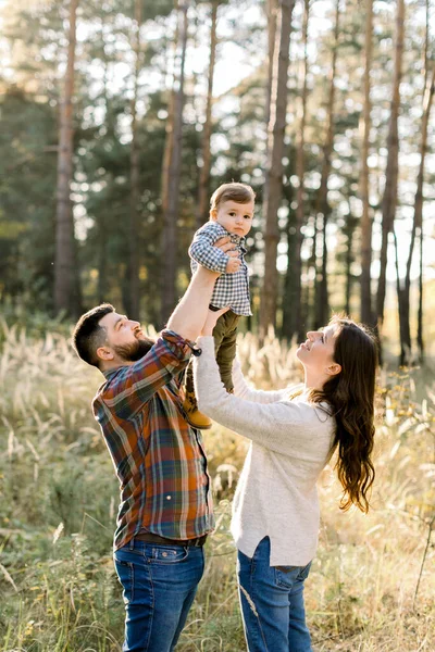 Güneşli bir günde, sonbahar ormanlarında yürüyüş yaparken şık gündelik giysiler giyen, eğlenen ve küçük tatlı oğullarını kaldıran mutlu aile portresi. — Stok fotoğraf