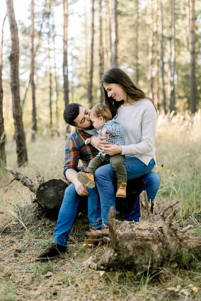 Açık havada mutlu beyaz bir ailenin portresi, baba, anne ve küçük bebek, şık günlük kıyafetler giyiyorlar, çam ağacındaki bir kütükte oturuyorlar ve kameraya gülümsüyorlar. — Stok fotoğraf