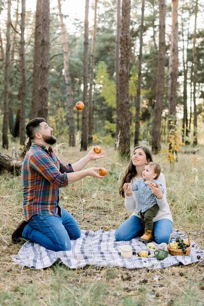 Familie, plezier en vrije tijd concept. Portret van een gelukkig gezin zittend op geruite ruitjes in het herfstbos. mooie mam en kleine baby jongen hebben plezier en kijken naar knappe vader, jongleren appels — Stockfoto