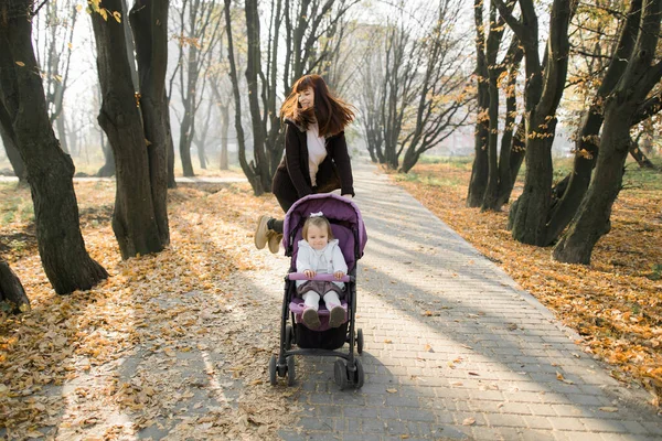 Οριζόντια εξωτερική φωτογραφία της ευτυχούς ελκυστική νεαρή μητέρα περπάτημα με το κοριτσάκι της, κάθεται σε ροζ καροτσάκι στο όμορφο πάρκο φθινόπωρο με πεσμένα κίτρινα φύλλα. Ευτυχής γονική αντίληψη — Φωτογραφία Αρχείου