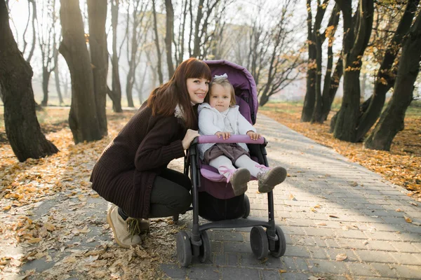 Happy mother in casual warm sweater, walking with her cute little daughter sitting in pink pram, in beautiful autumn park with yellow fallen leaves at sunny day, smiling and posing at camera. — Stock Photo, Image