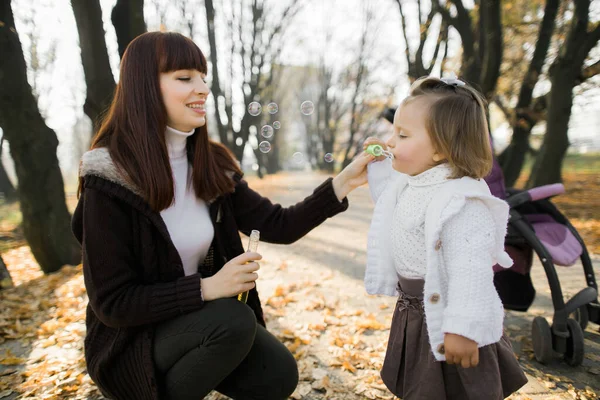 Close up retrato de mãe feliz e bebê menina criança, se divertindo no belo parque de outono no dia ensolarado, jogando, sorrindo, menina litlle está soprando bolhas de sabão — Fotografia de Stock
