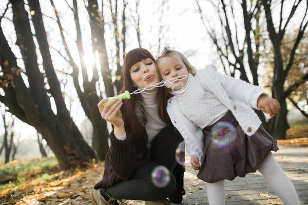 Horizontal outono brilhante tiro de mãe muito jovem mulher, se divertindo e soprando bolhas de sabão, juntamente com sua filhinha bonito, andando no dia ensolarado no parque de outono — Fotografia de Stock