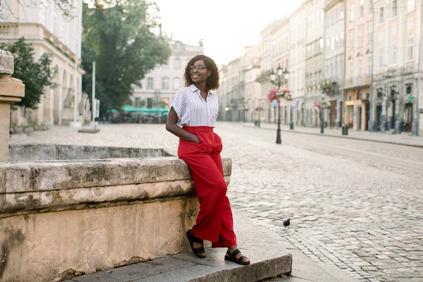 Outdoor full length portret van jonge mooie Afrikaanse zakenvrouw, het dragen van rode broek en gestreept shirt, rusten na harde dag, poseren in het oude Europese centrum, in de buurt van stenen monument op trappen — Stockfoto