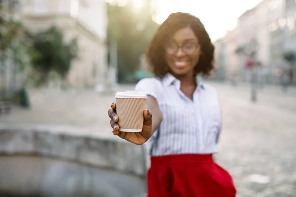 Afhaalkoffie in de stad buiten. Glimlachende jonge Afrikaanse vrouw in rode broek en wit shirt, tonen aan de camera papieren beker met warme drank, staan op straat buiten. Focus op de kop koffie — Stockfoto