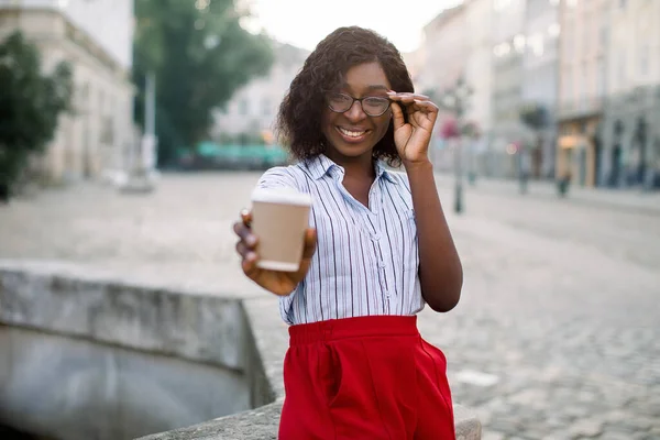 Jonge elegante Afrikaanse zakenvrouw in slimme casual kleding, het houden van afhaalkoffie kopje tijdens het lopen buiten kantoor tijdens de pauze, glimlachend naar de camera. wazig oude stad op de achtergrond — Stockfoto