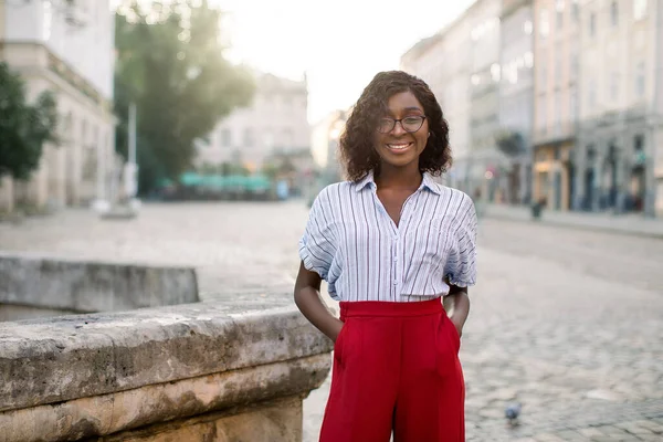 Portret van een glimlachende, vrolijke, modieuze Afrikaanse vrouw met een bril, een rode broek en een gestreept shirt op de oude straat van de stad. Oude gebouwen, ochtendstad wazig achtergrond — Stockfoto