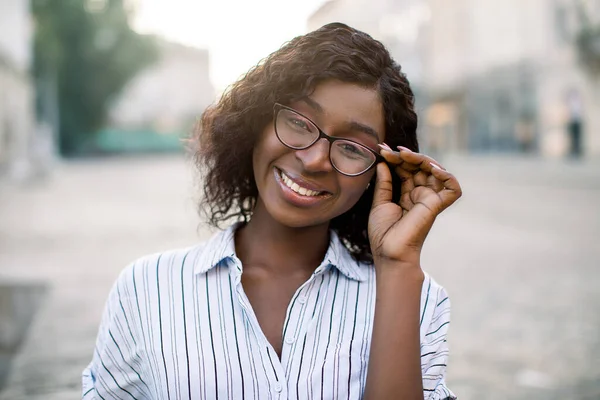 Outdoor stedelijke close-up portret van stijlvolle positieve Afrikaanse vrouw in gestreept casual shirt, aanraken van haar bril, glimlachen om camera wandelen buiten, op wazig stad straat achtergrond — Stockfoto