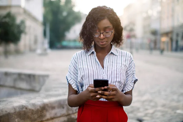 Close-up horizontaal portret van aantrekkelijke jonge Afrikaanse vrouw, in rode broek en gestreept shirt, lezen of typen sms-bericht op mobiele telefoon in de oude stad straat. Mensen, technologie concept — Stockfoto