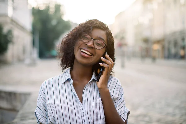 Close-up portret van aantrekkelijke krullend Afrikaans meisje praten op mobiele telefoon, kijken naar camera en lachen, wandelen buiten op de oude stad straat bij zonsondergang. Mensen, stedelijke stijl, technologie concept — Stockfoto